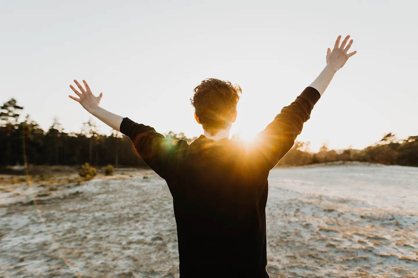 Auf diesem Bild zu sehen ist ein Mann von hinten, der in der Natur steht. Die Sonne geht gerade auf, er begrüßt sie mit hochgestreckten Armen. 