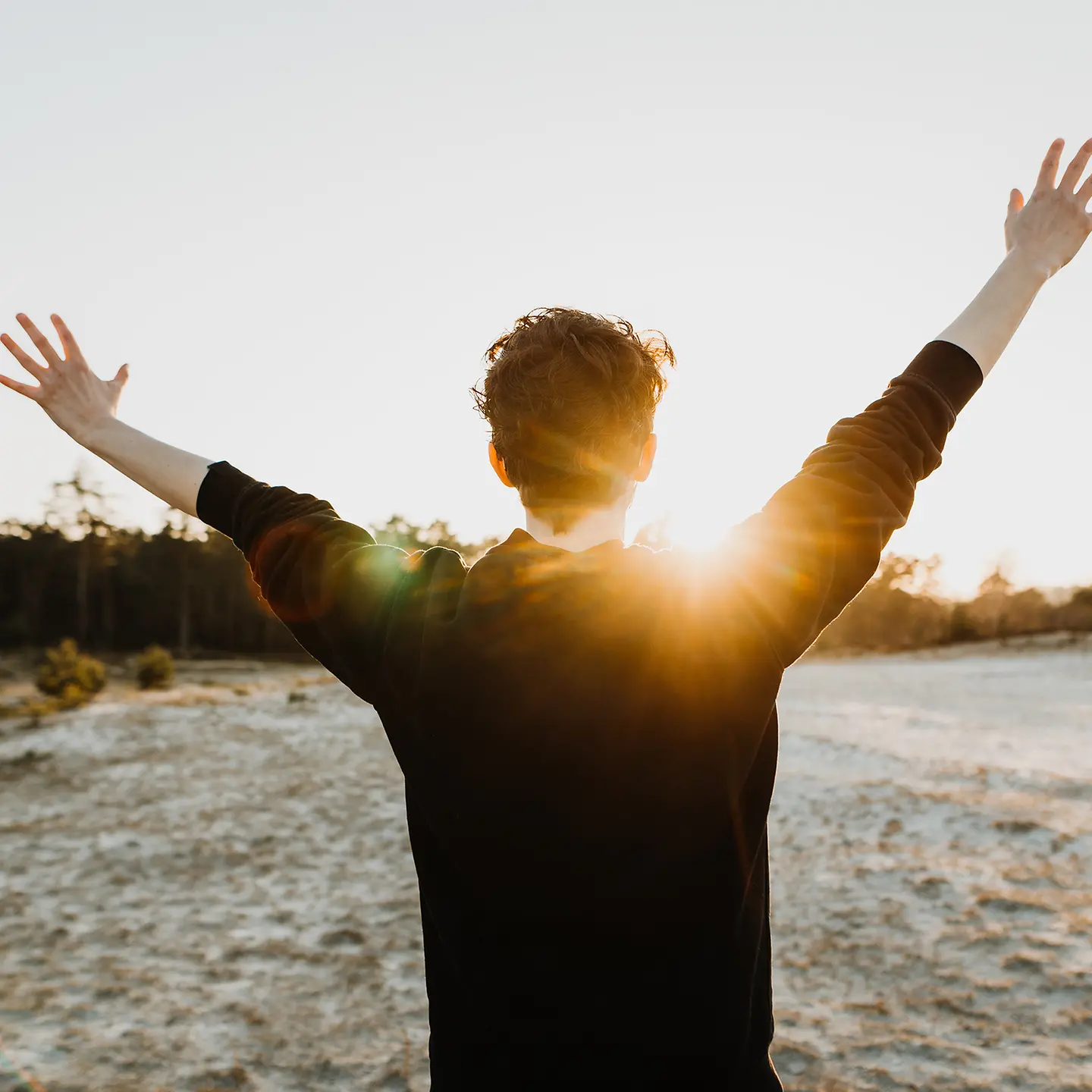 Auf diesem Bild zu sehen ist ein Mann von hinten, der in der Natur steht. Die Sonne geht gerade auf, er begrüßt sie mit hochgestreckten Armen. 