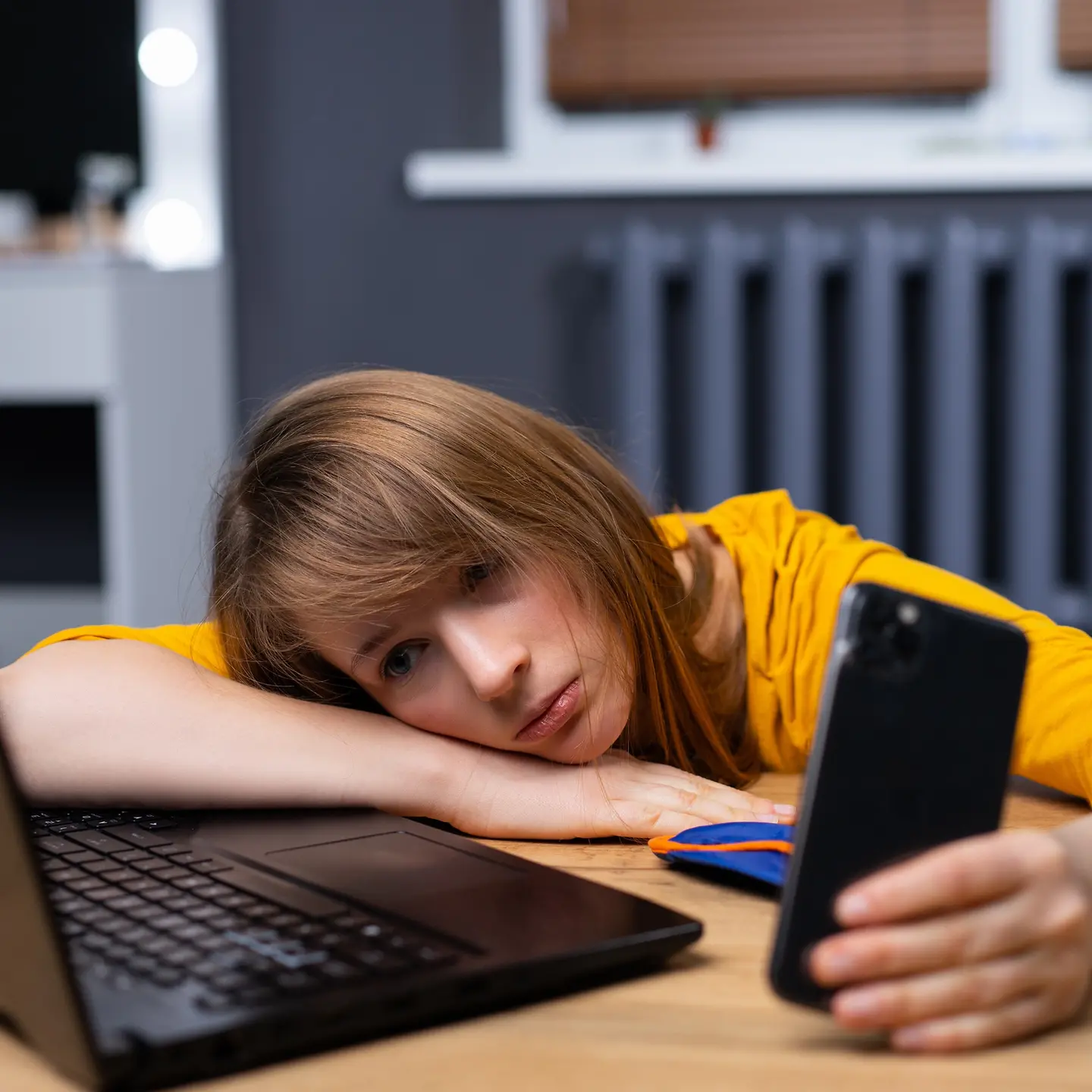 Auf dem Bild zu sehen ist eine überarbeitete, müde Frau: eine junge, erschöpfte Studentin im Büro. Die Frau nutzt Laptop und Smartphone im Homeoffice.