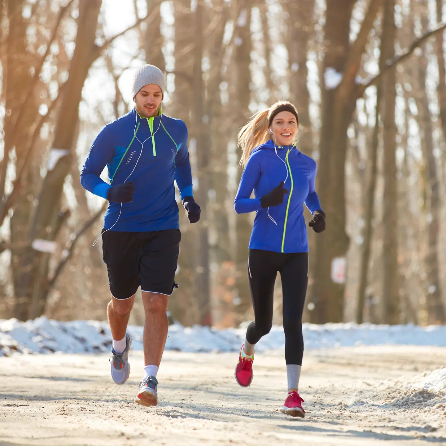 uf dem Bild ist ein junges Paar zu sehen, die im Schnee im winterlichen Wald joggen oder laufen gehen. Die Frau lächelt in die Kamera.