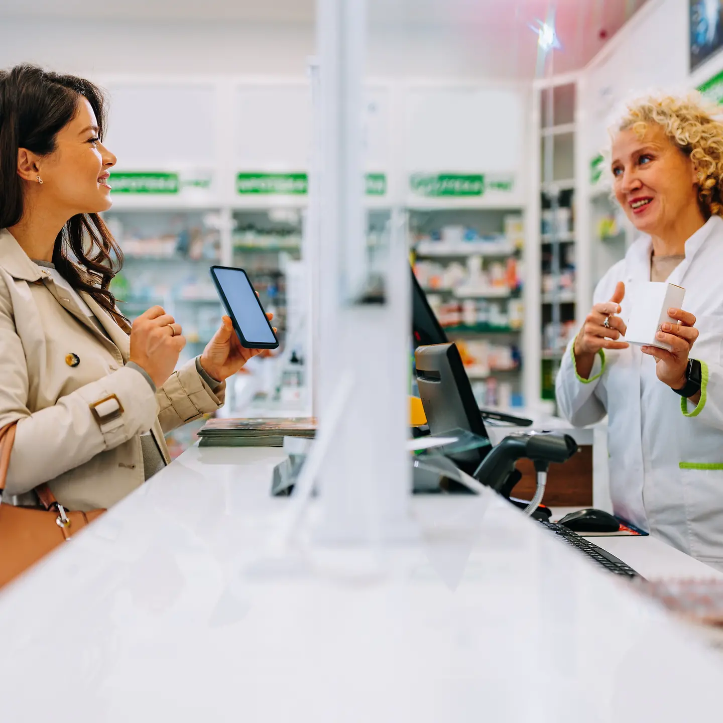 Auf dem Bild ist eine Frau zu sehen, die mit ihrem Handy in der Apotheke Medikamente kauft, sie lächelt. Die Apothekerin hält ein Medikament in der Hand und lächelt auch.