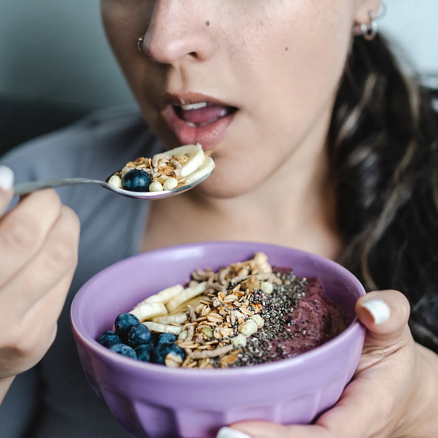 Auf dem Bild zu sehen ist junge Frau mit dunklen Haaren, die gerade Müsli isst. Es besteht aus Beeren, Körnern, Bananen und sieht sehr gesund aus.