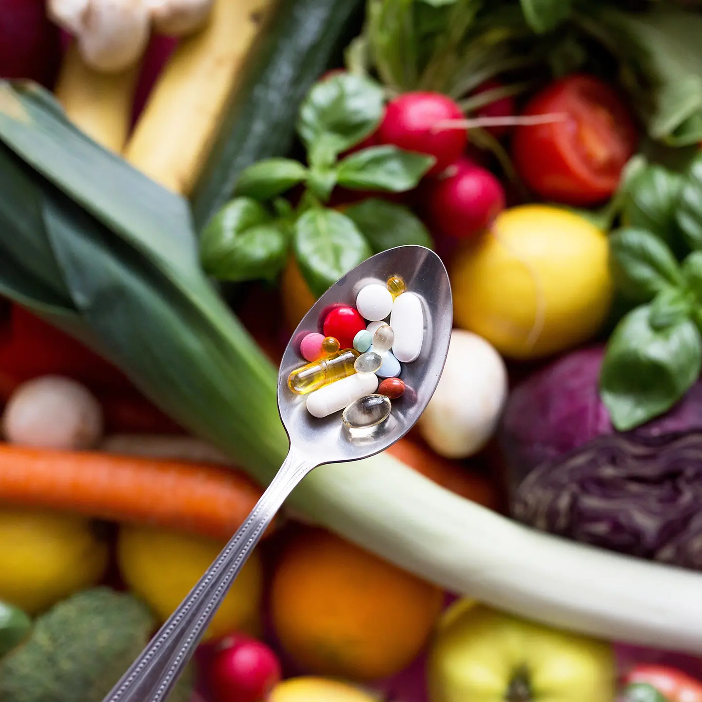 Auf dem Bild sind im Hintergrund leicht unscharf Gemüse und Obst zu sehen. Den Mittelpunkt bildet scharf ein Löffel mit Nahrungsergänzungsmittel.