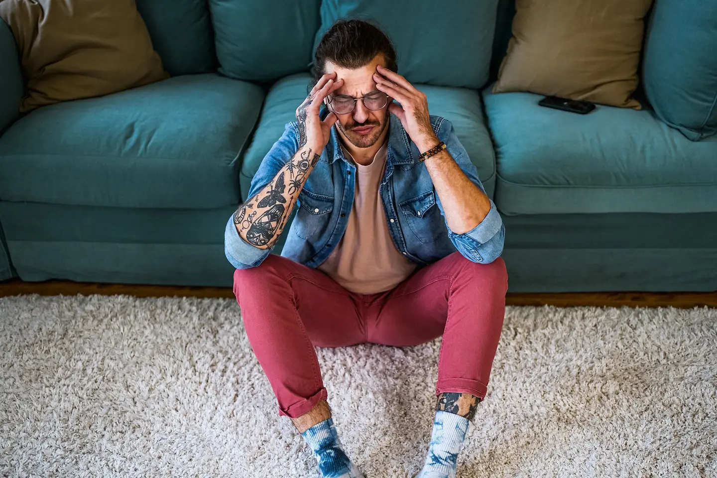 Mann sitzt vor einem auf Sofa mit Kopfschmerzen und hält sich die Hände an den Kopf. 