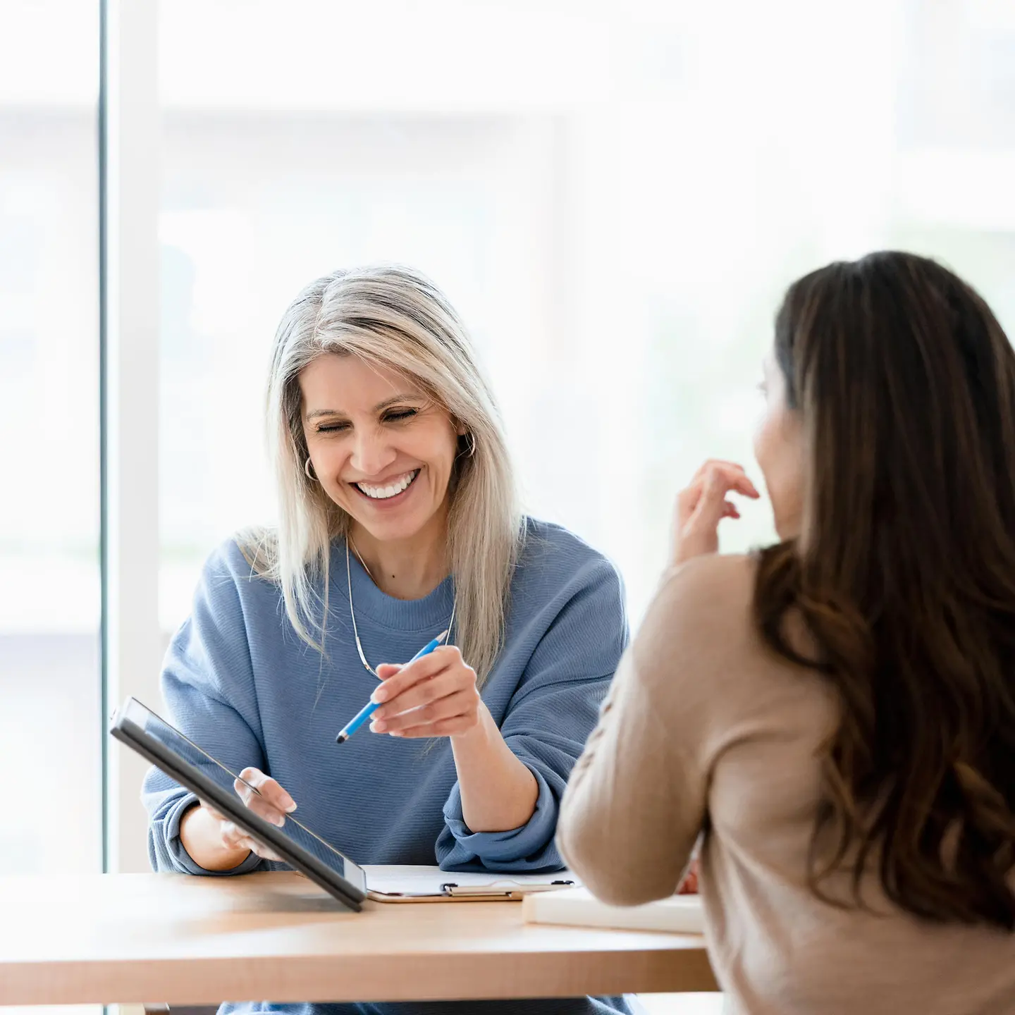 Zwei Frauen sitzen in einem Büro gemeinsam an einem Tisch und schauen zusammen auf ein Tablet.