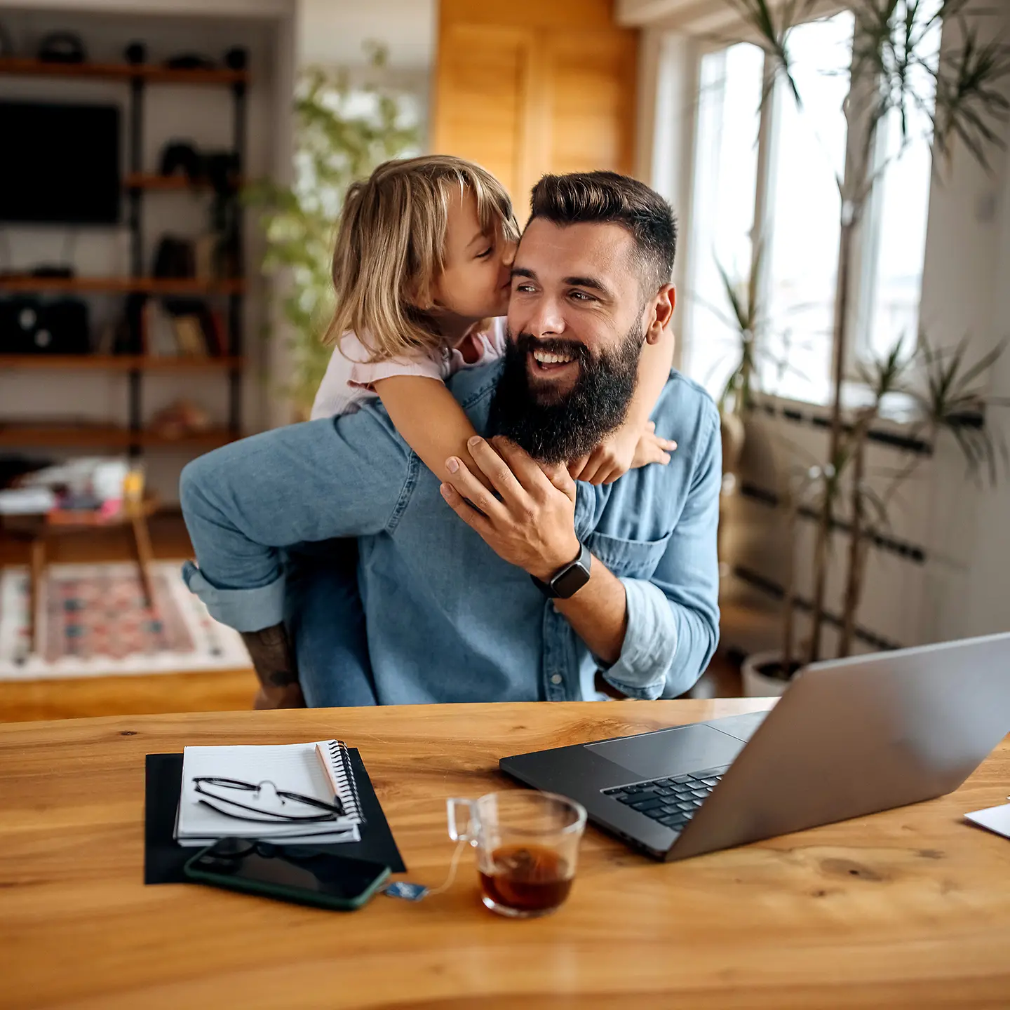 Ein Mann arbeitet im Homeoffice mit einem Laptop am Tisch, während ein kleines Kind ihm lächelnd von hinten um den Hals fällt.