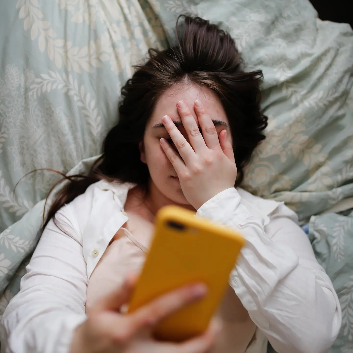 Junge Frau liegt im Bett, hält ihr Handy und hat ihr Gesicht mit der Hand verdeckt