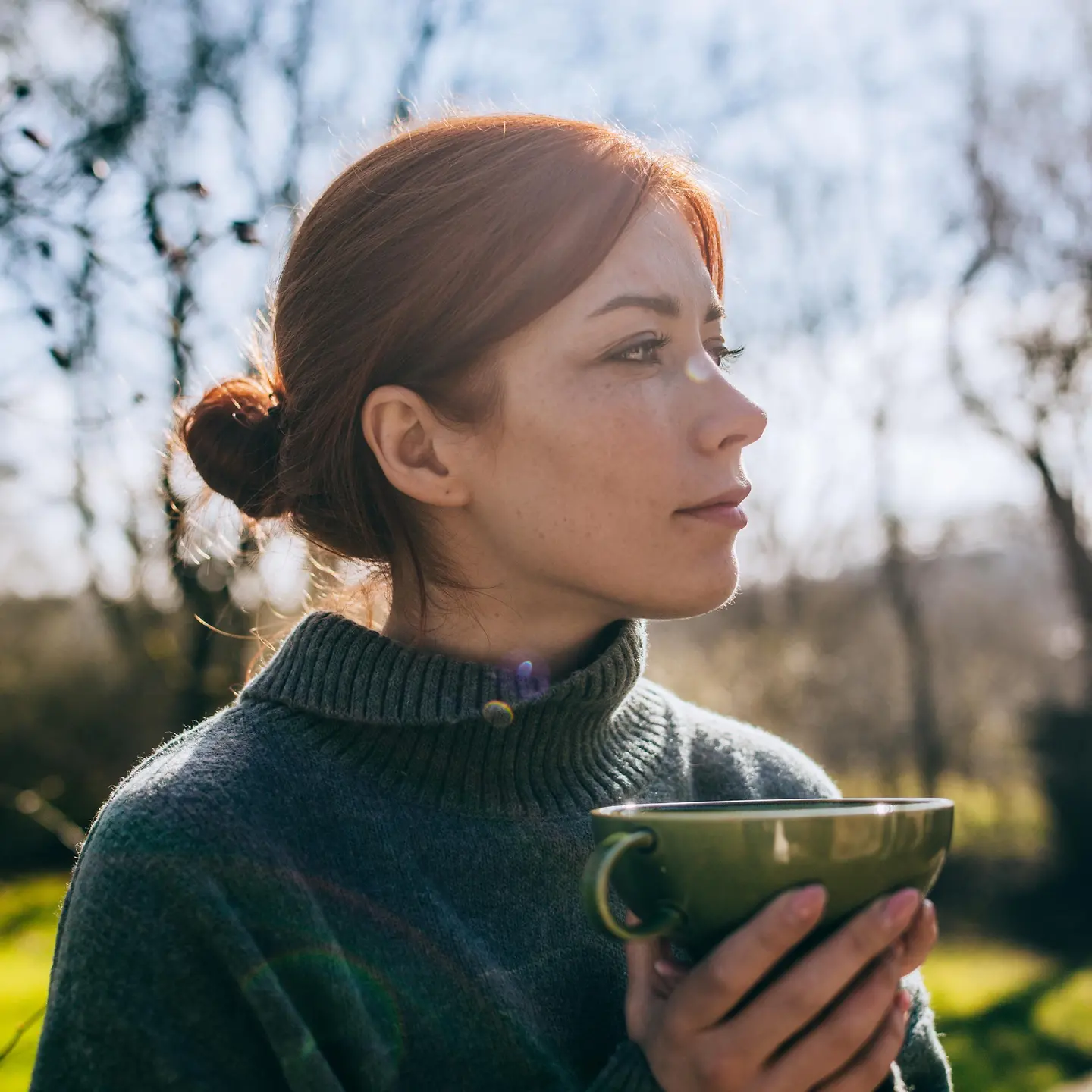 Eine junge Frau steht im Garten mit einer Tasse in der Hand und schaut nachdenklich