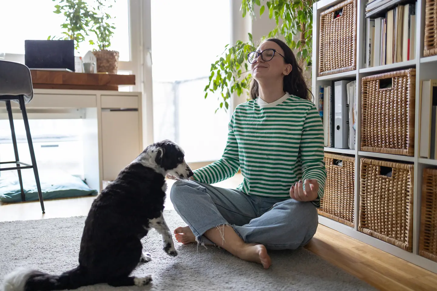Eine junge Frau meditiert zu Hause. Ein Hund sitzt vor ihr.