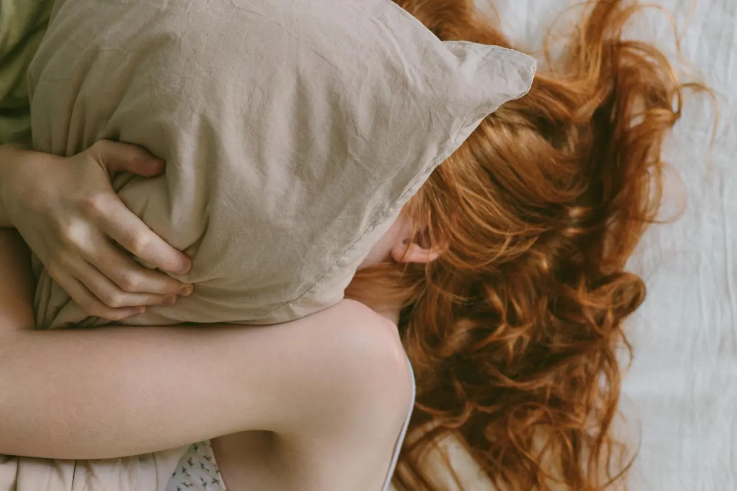 Junge Frau mit roten Haaren liegt im Bett unter dem Kopfkissen versteckt