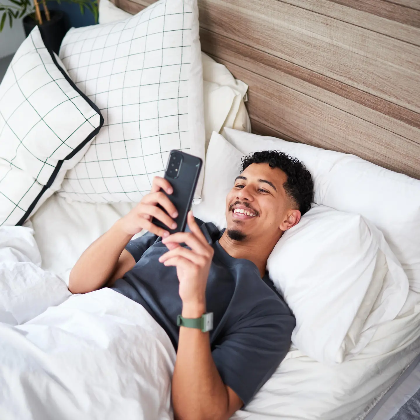 Junger Mann liegt im Bett und schaut lächelnd auf sein Handy