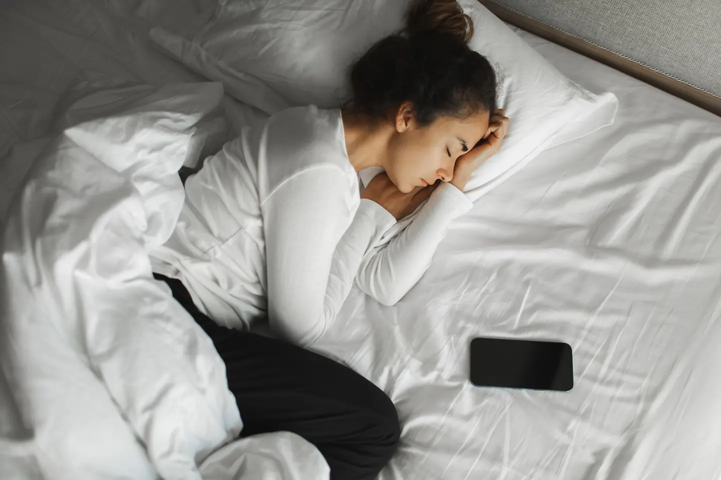 Junge Frau liegt schlafend im Bett mit einem Handy neben sich