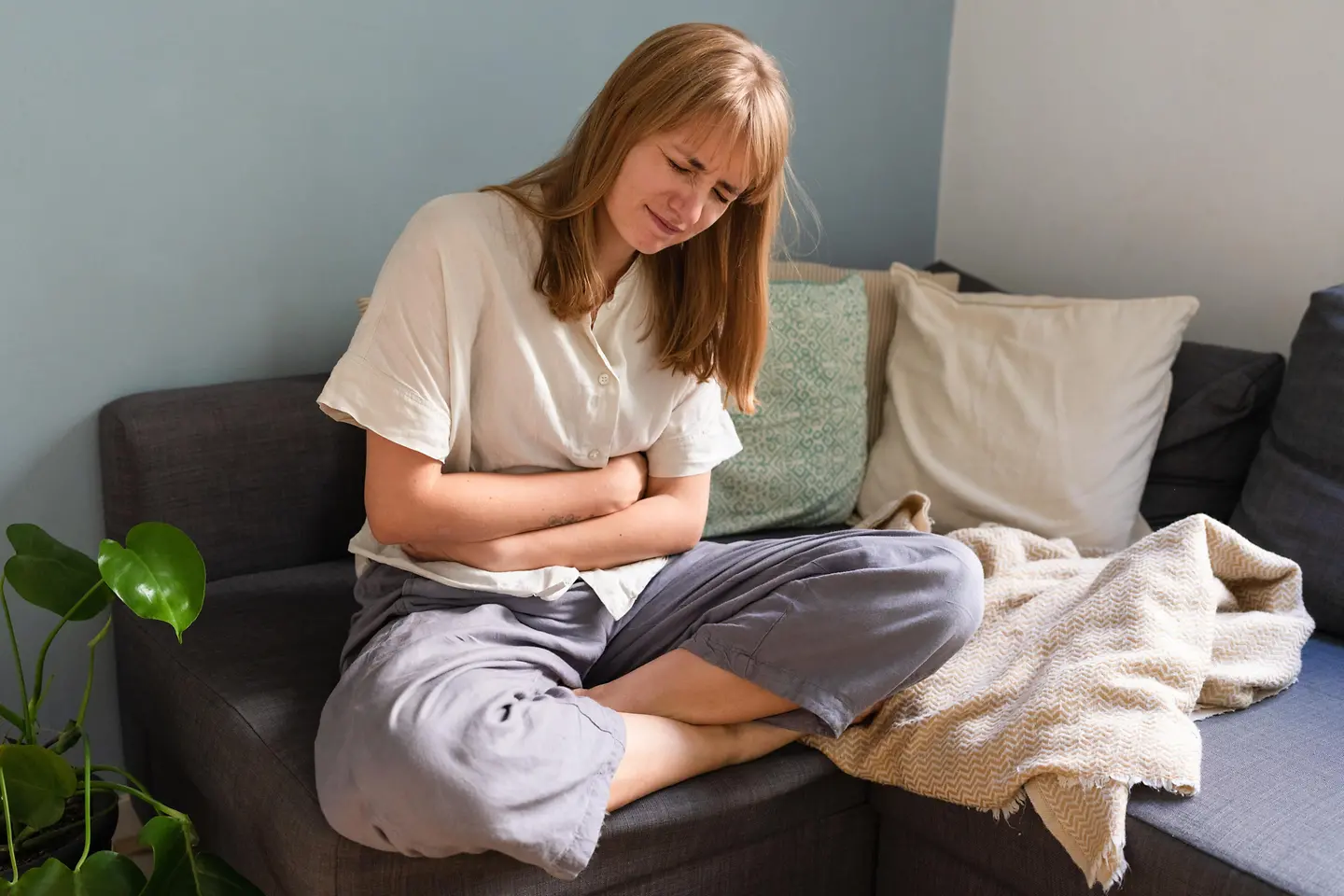 Eine Frau sitzt gekrümmt mit Bauchschmerzen auf dem Sofa und verschränkt die Arme vor dem Bauch.