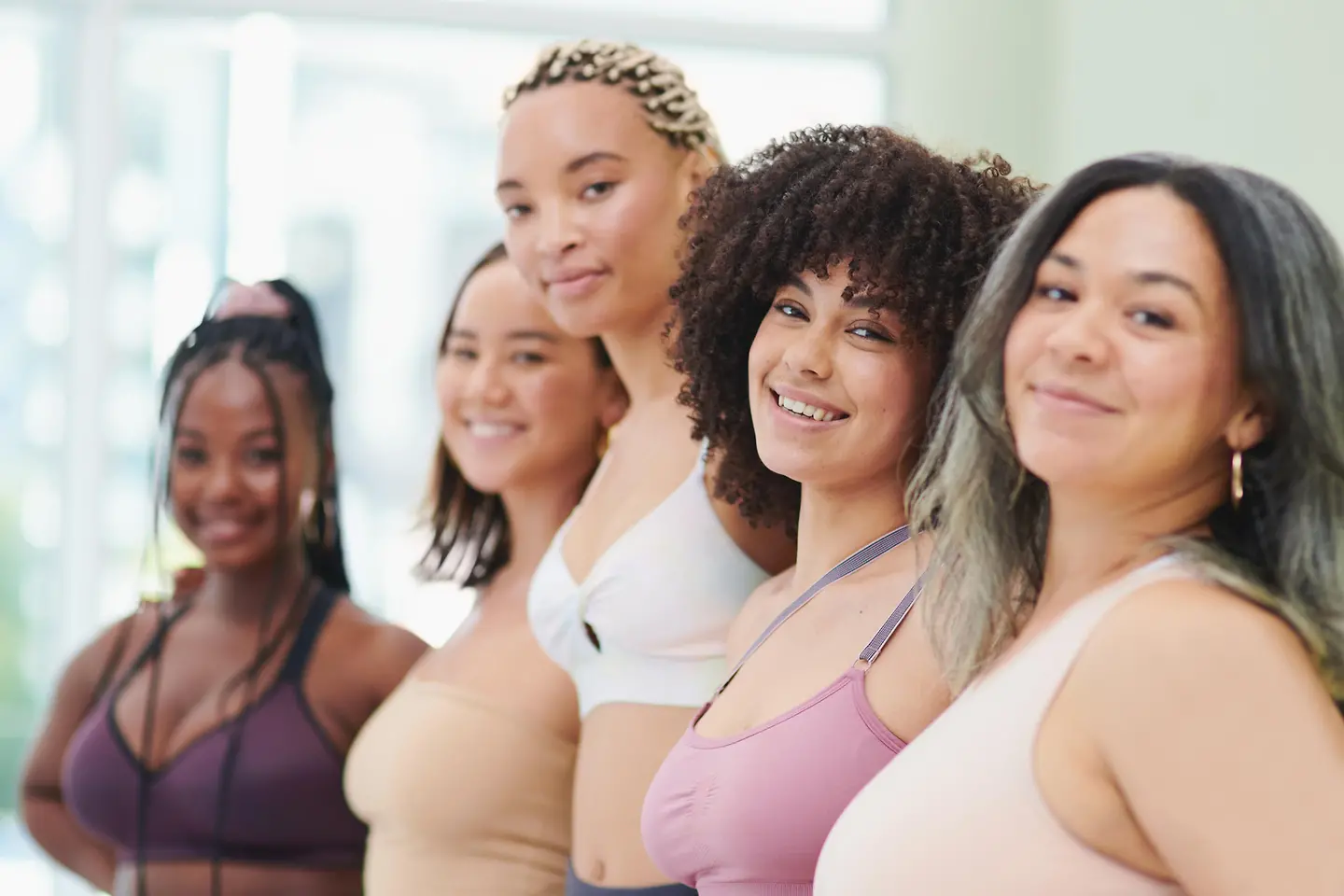 Gruppe von Frauen in Fitnesskleidung, die vor einem hellen, unscharfen Hintergrund zusammenstehen und lächeln