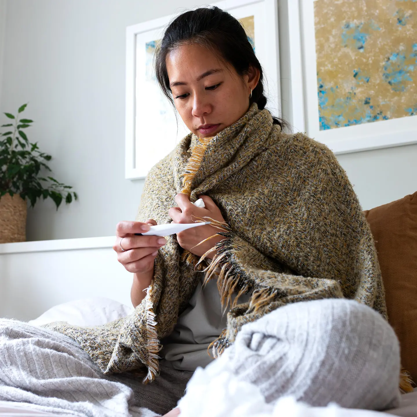 Frau sitzt im Bett in eine Decke eingewickelt und schaut auf ein Fieberthermometer
