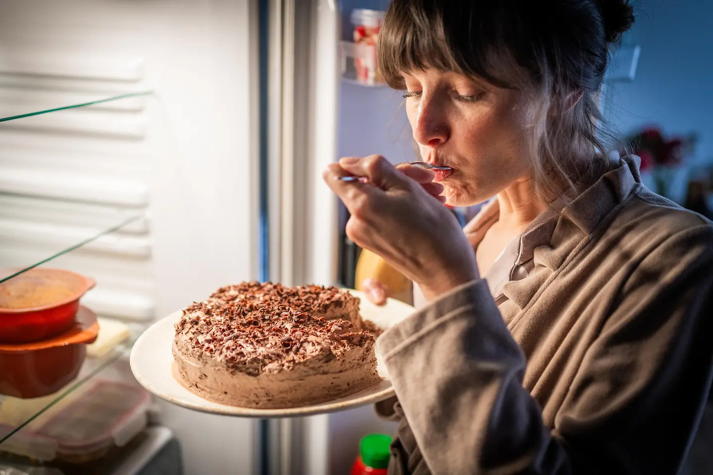 Junge Frau steht nachts am Kühlschrank und isst heimlich Schokotorte
