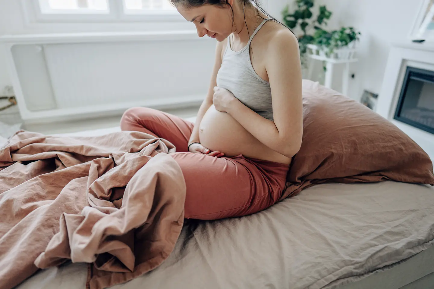 Eine junge schwangere Frau sitzt auf dem Bett und streichelt ihren Babybauch