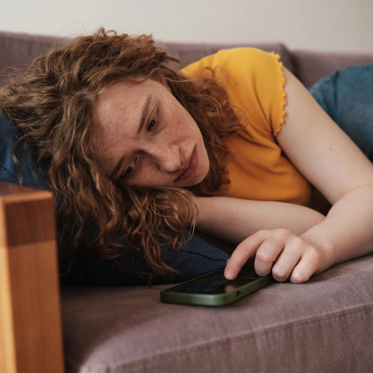 Junge Frau mit roten Haaren liegt auf einer Couch und schaut auf ihr Handy