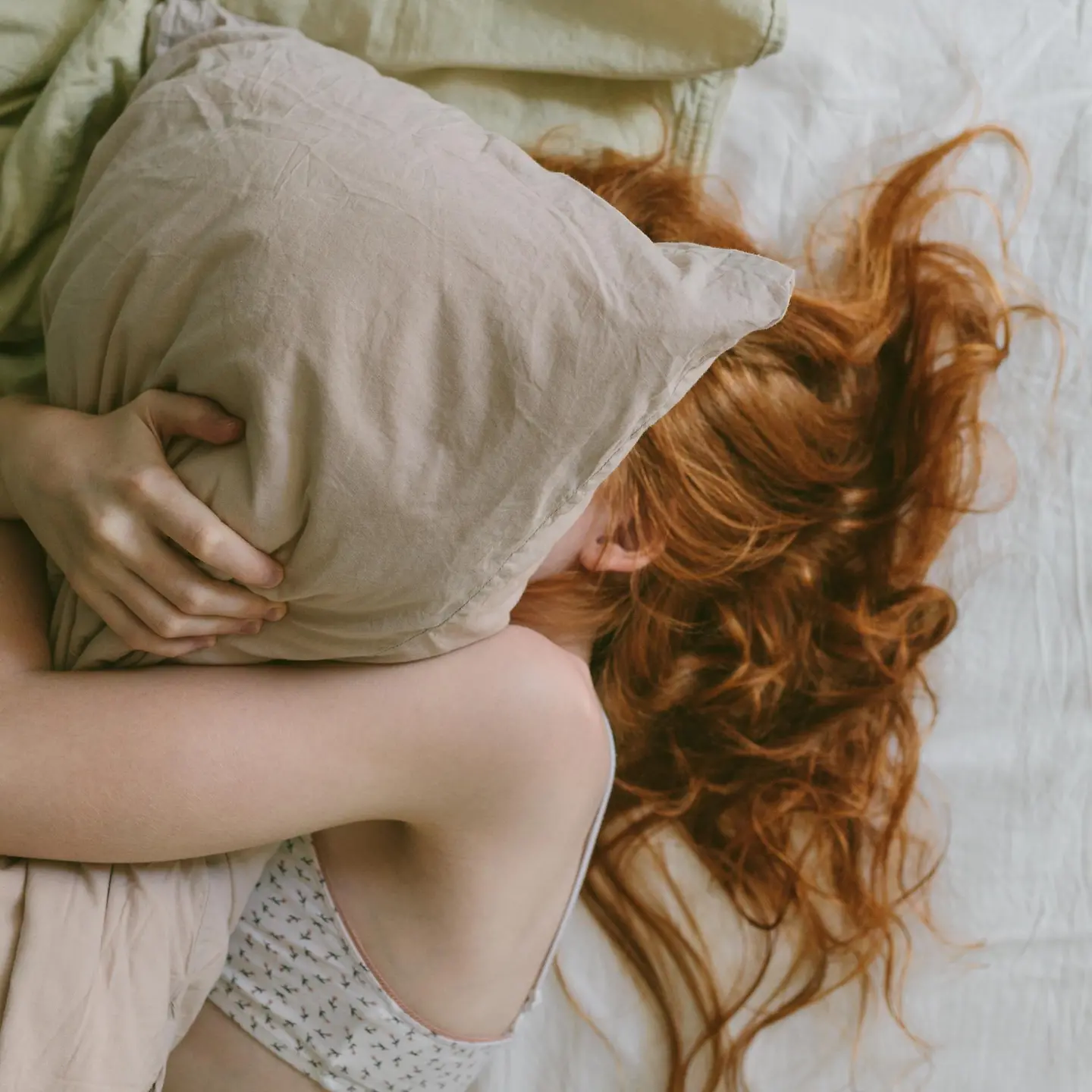Junge Frau mit roten Haaren liegt im Bett unter dem Kopfkissen versteckt