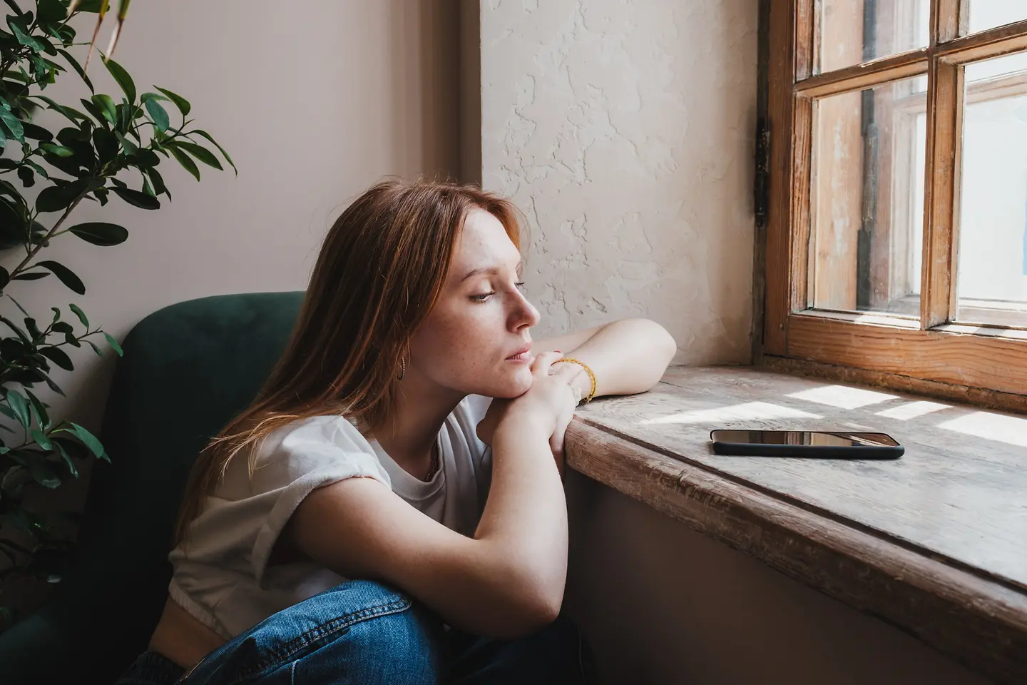 Eine junge Frau sitzt an einem Fenster und schaut nachdenklich auf ihr Smartphone