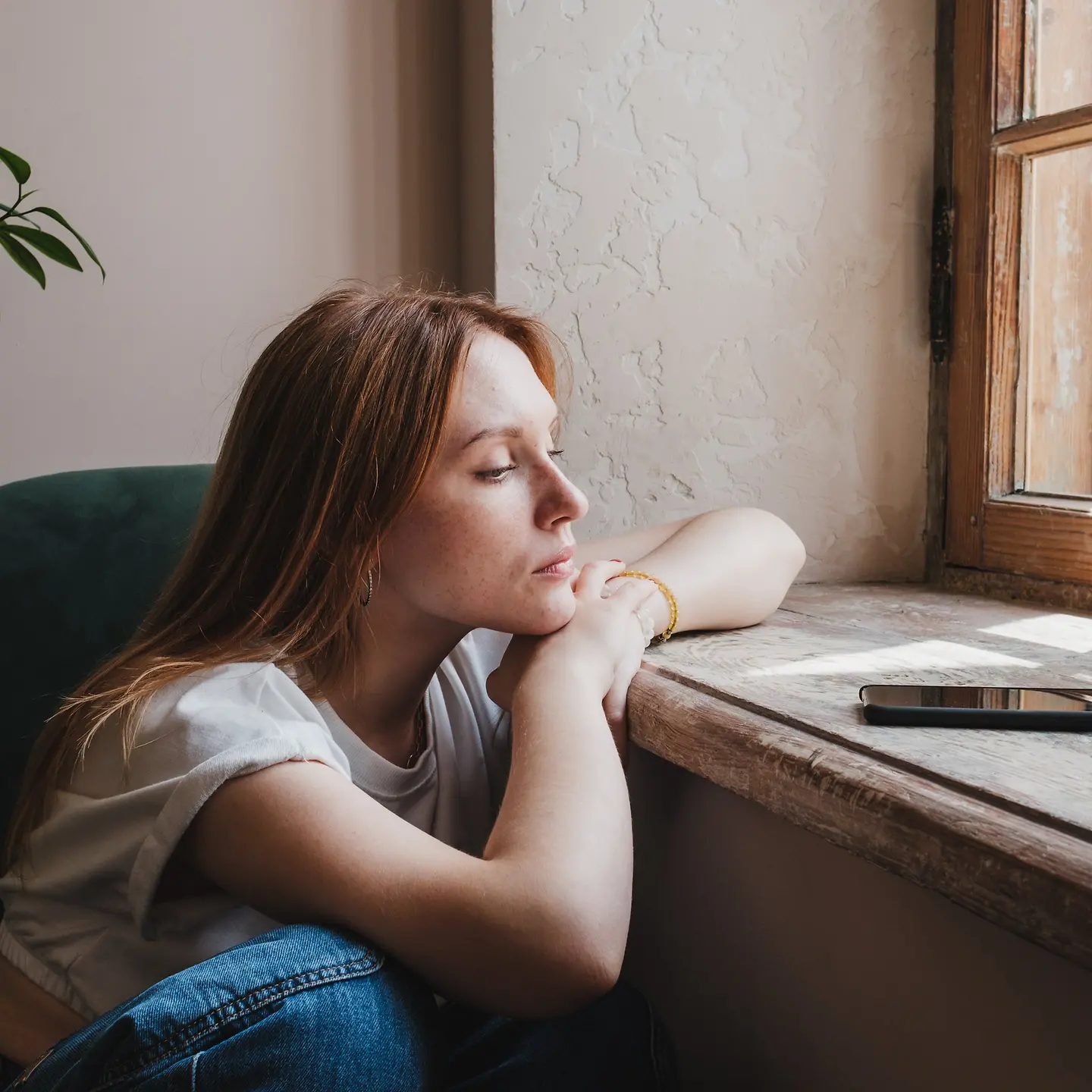 Eine junge Frau sitzt an einem Fenster und schaut nachdenklich auf ihr Smartphone