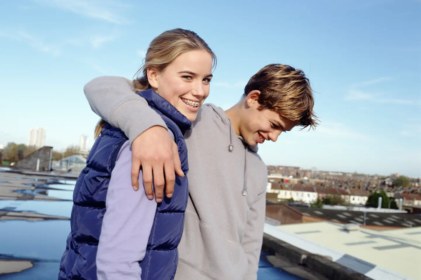 Ein Teenager-Paar (14-16) steht auf einem Dach mit Blick über die Stadt. Der Junge hat seinen um das Mädchen gelegt. Beide lächeln.