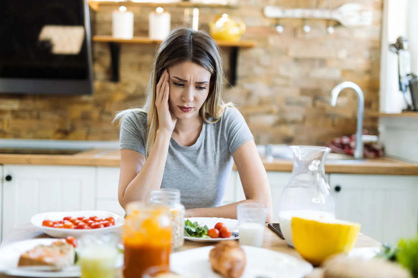 Traurig schauende junge Frau sitzt am Küchentisch und will ihr gesundes Frühstück mit Tomaten und Melone nicht essen