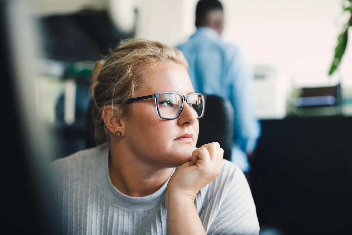 Eine Frau sitzt nachdenklich im Büro, stützt das Kinn auf die Hand und blickt in die Ferne.