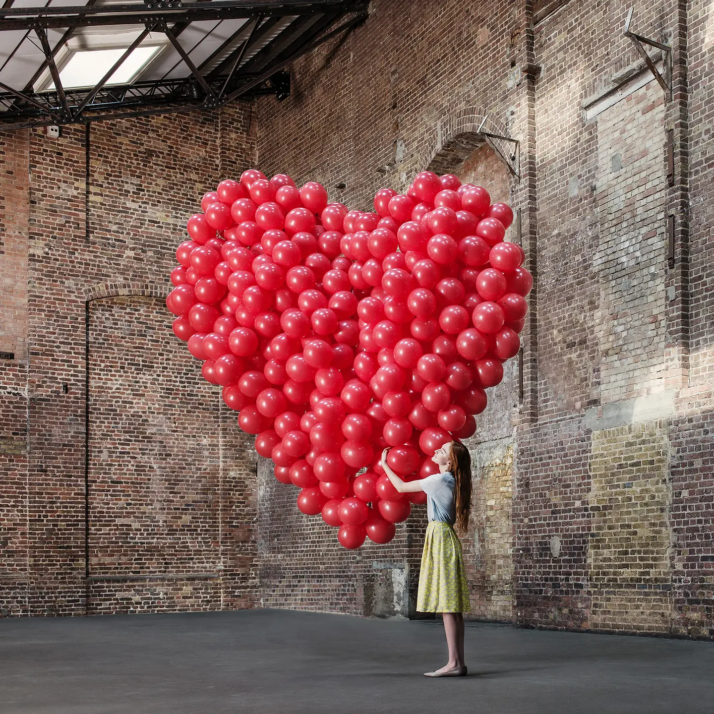 Selbstliebe: Junge Frau steht in einer Halle und umarmt ein grosses Herz aus roten Luftballons