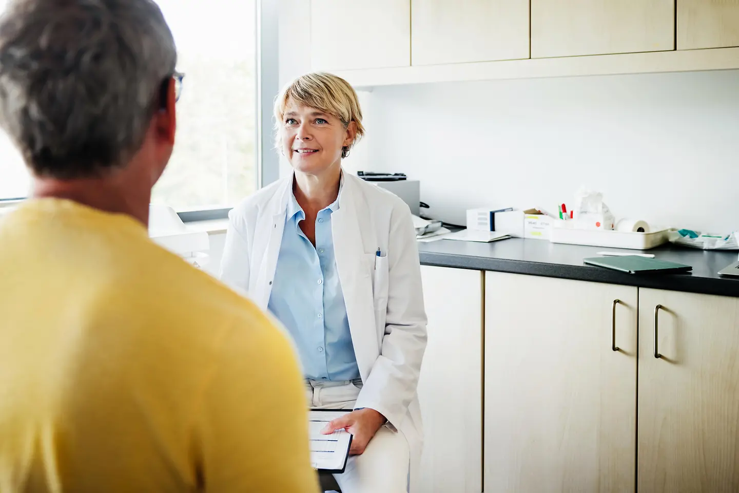 Eine Ärztin im weißen Kittel spricht in einem hellen Behandlungszimmer mit einem Patienten, der mit dem Rücken zur Kamera sitzt.
