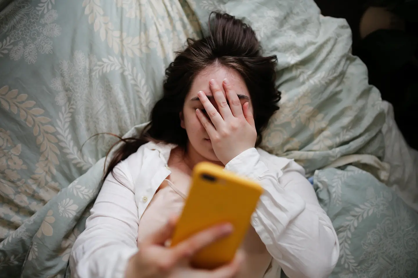 Junge Frau liegt im Bett, hält ihr Handy und hat ihr Gesicht mit der Hand verdeckt