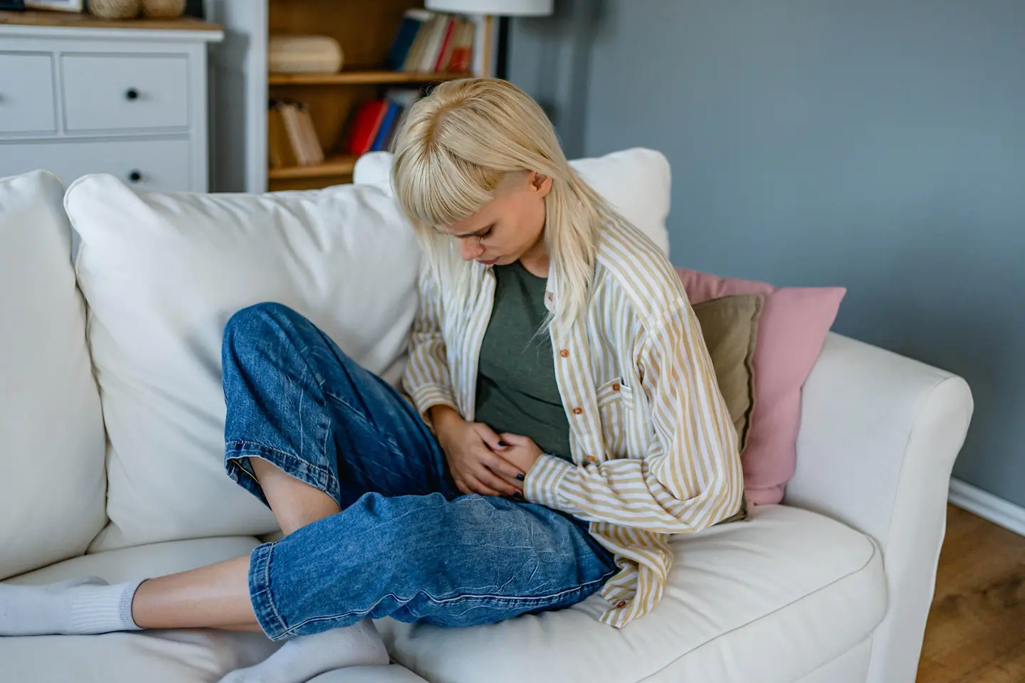 Eine Frau sitzt zusammengekauert auf einem Sofa und hält beide Hände auf ihren Bauch. Symbolbild für Verstopfung.